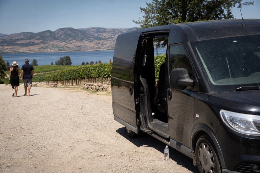 A private van parked at a winery entrance with vineyard slopes and lake water beyond, no text, no logos, no people