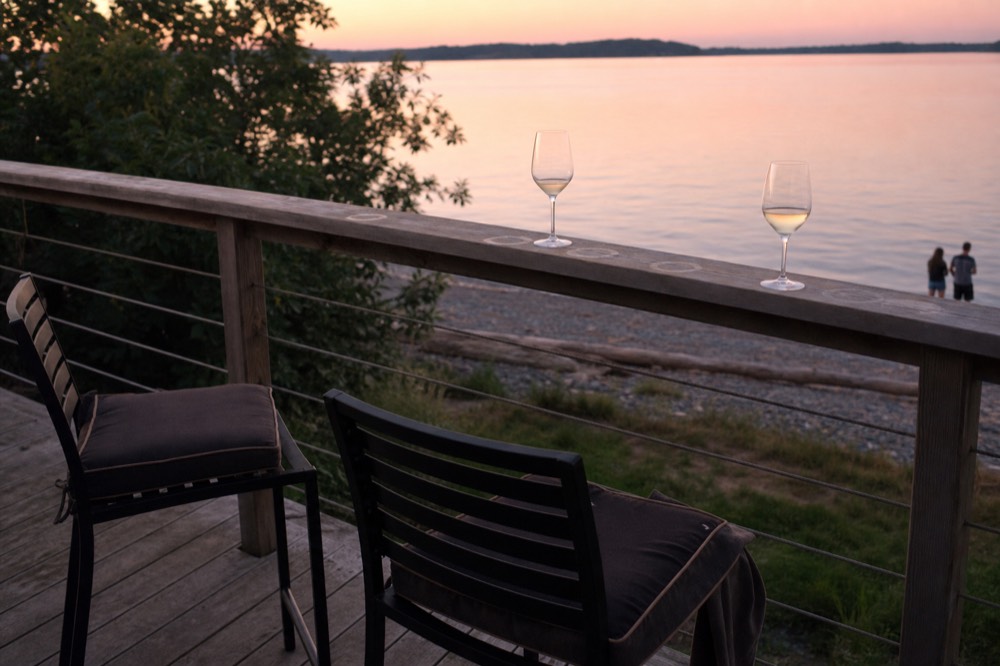 A tasting room deck overlooking calm water with chairs and glasses on a rail, no text, no logos, no people