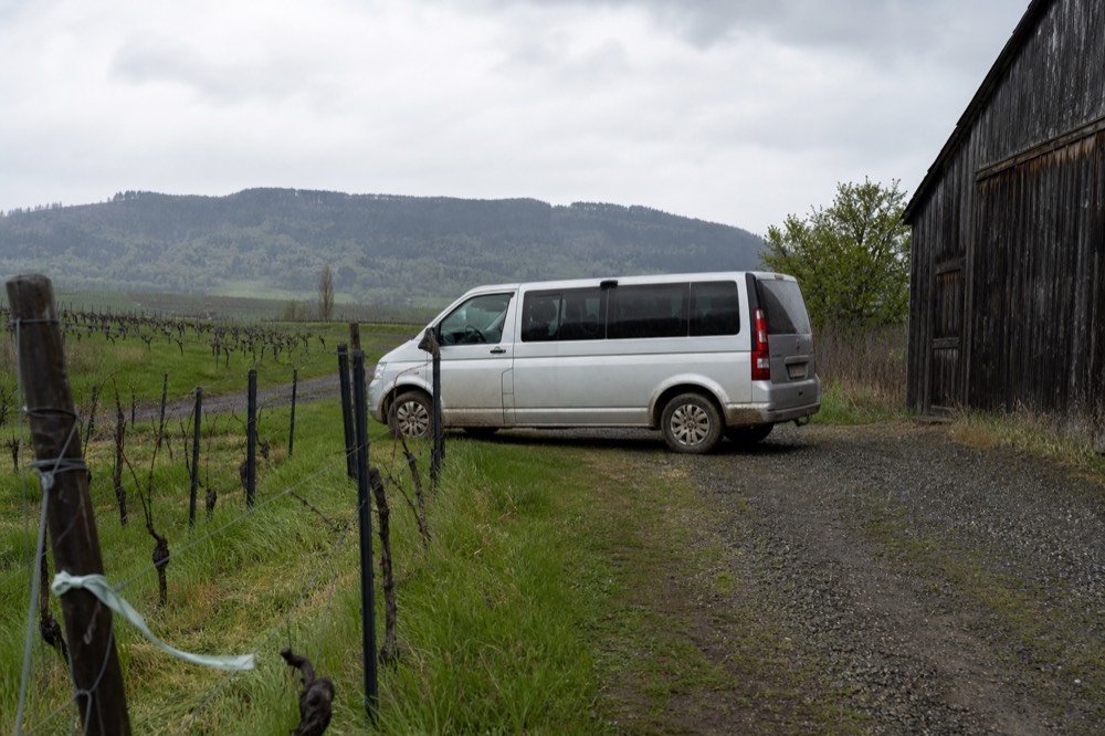 A small van parked on a rural road between vineyards and a barn, no text, no logos, no people