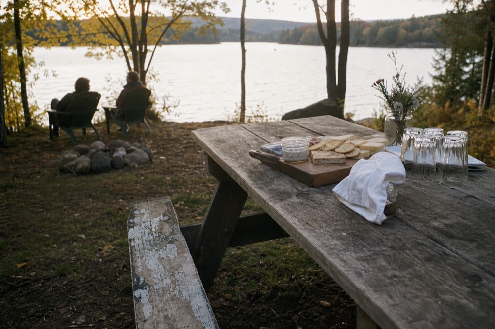 A picnic table with a cheese board, crackers, and unopened wine glasses overlooking a lake, no text, no logos, no people