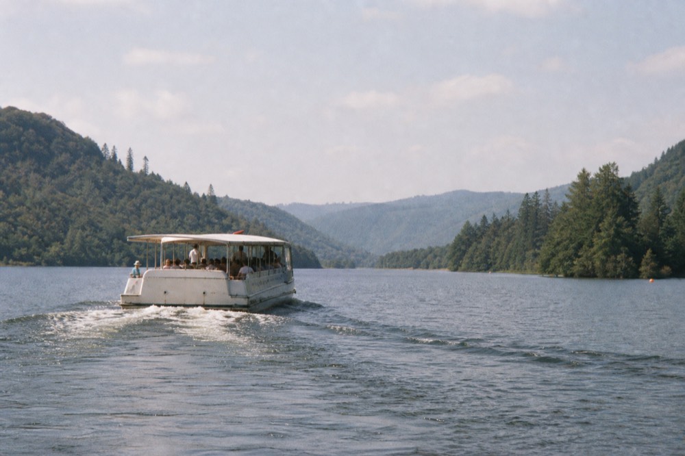 A tour boat crossing open water on a long glacial lake, forested hills rising on both shores, no text, no logos, no people