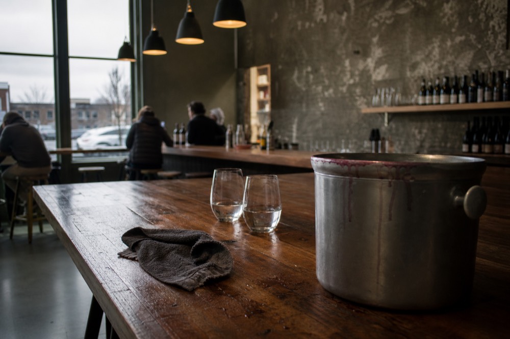 A dump bucket and water glasses on a wooden tasting bar, no text, no logos, no people.